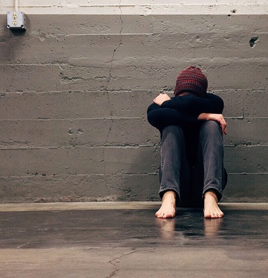 A young person sitting alone against a wall with their head resting on their arms